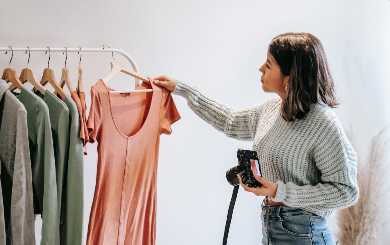 heros-img Young woman holding a camera while checking clothes on a rack indoors.
